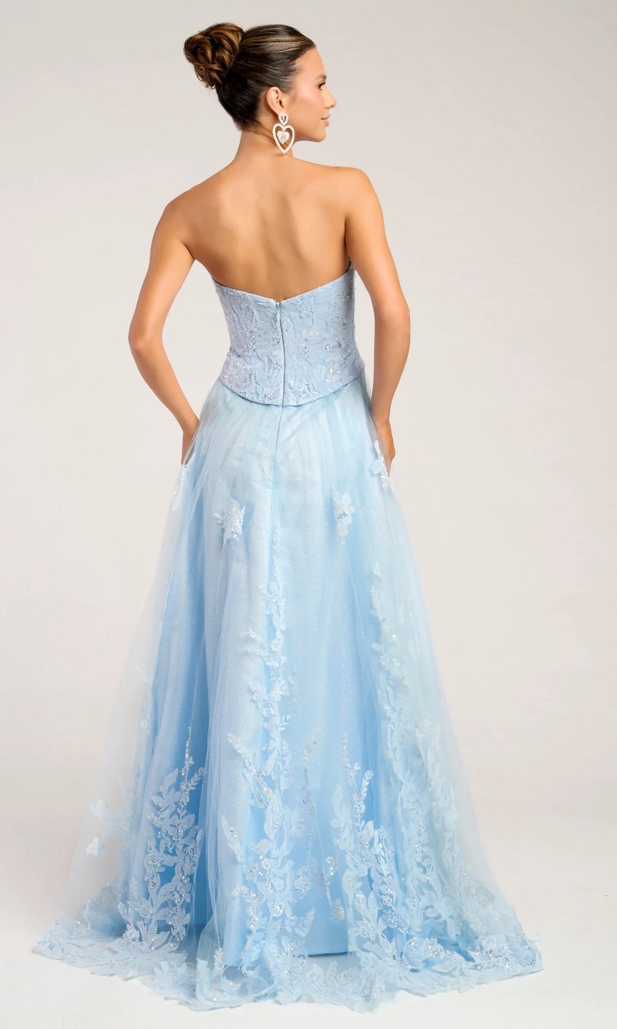 A woman faces away, wearing the Portia and Scarlett PS26061 long formal dress with a corset bodice and light blue floral lace details, hair in a bun, heart-shaped earrings, against a plain background.